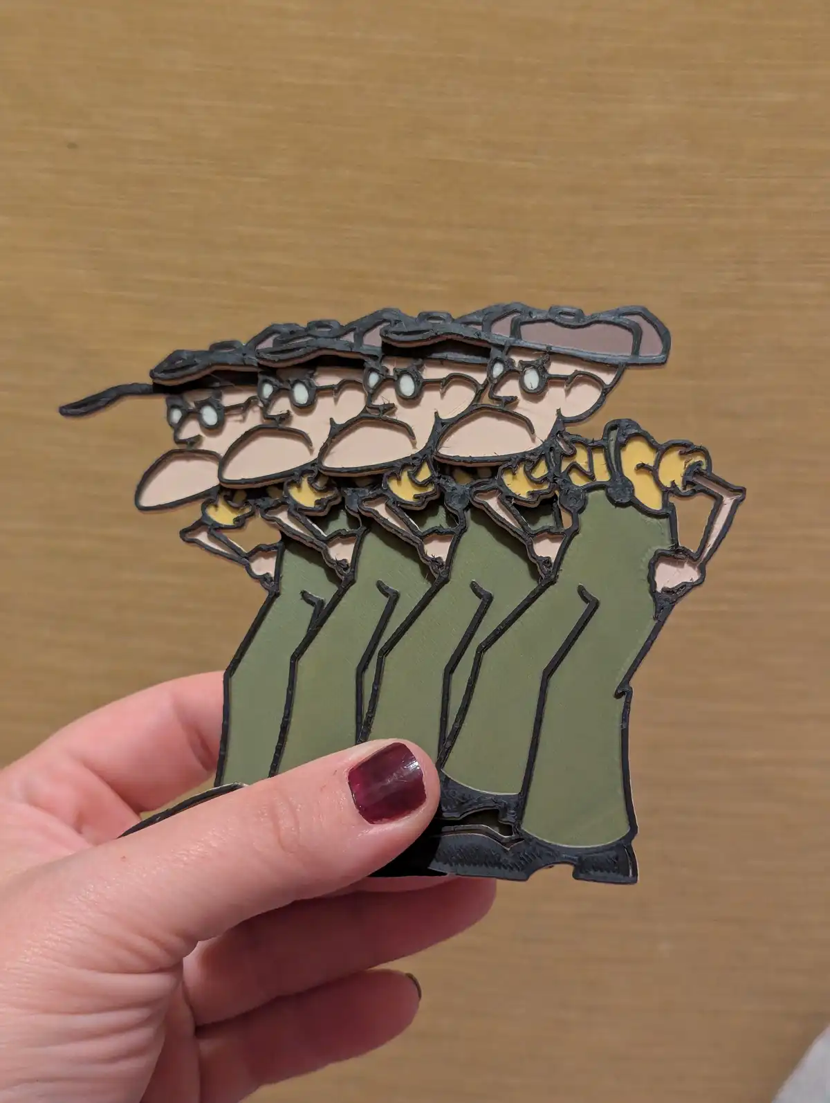 A hand holds a stack of identical 3D-printed cutout figures resembling soldiers in olive green uniforms, tan hats, and…