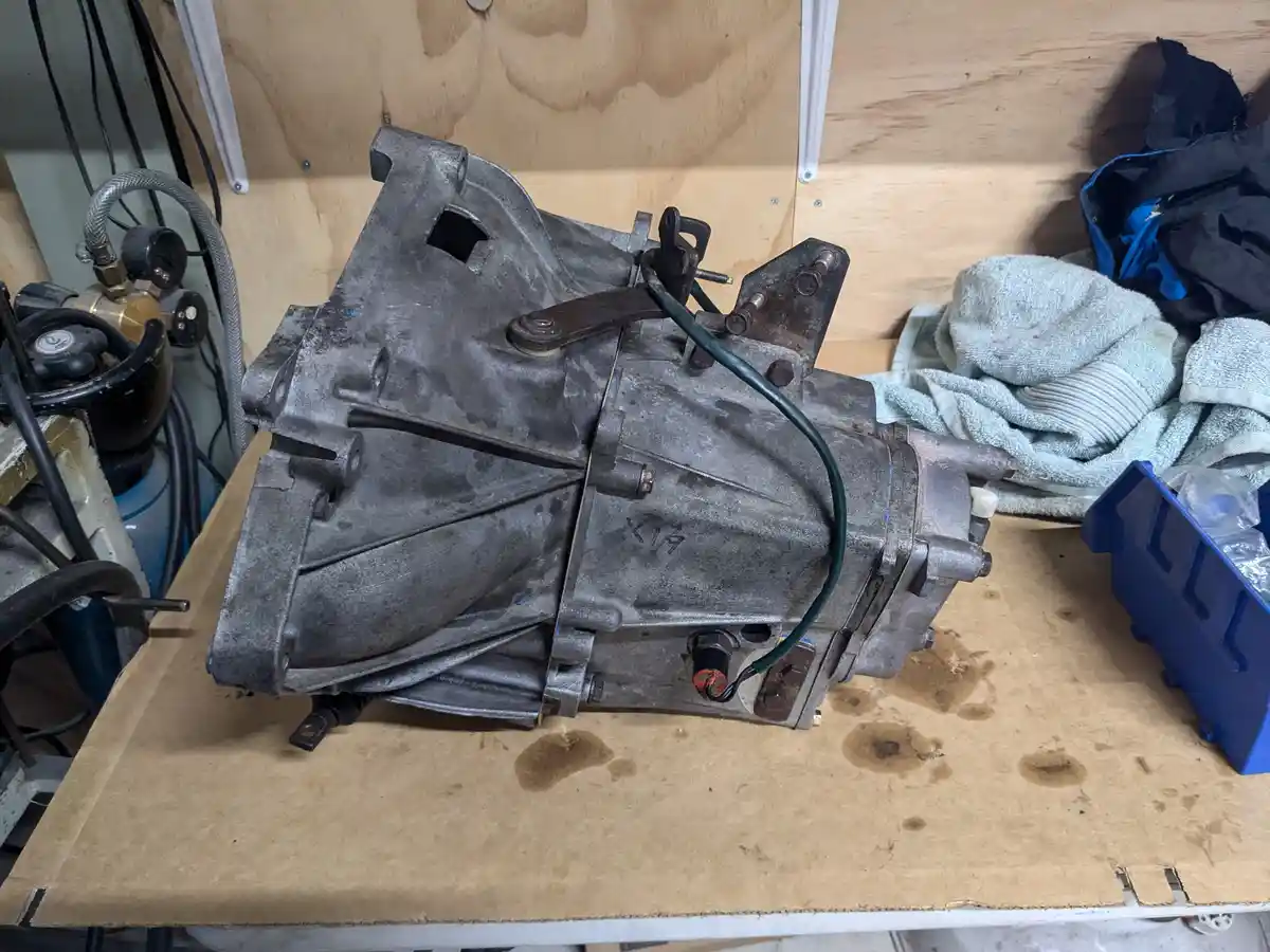 A dirty, metallic car transmission sits on a stained cardboard workbench in a garage, with tools and a blue toolbox nearby…