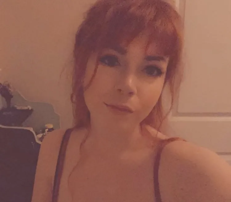 A selfie of a woman with reddish hair and bangs, wearing a dark tank top, looking directly at the camera with a neutral…