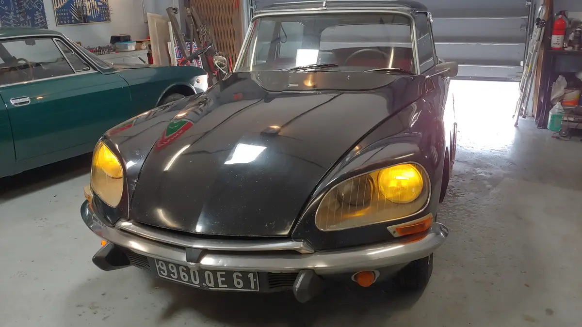 A black vintage Citroën DS car with its headlights on is parked in a garage, facing the camera. Behind it, a teal classic…