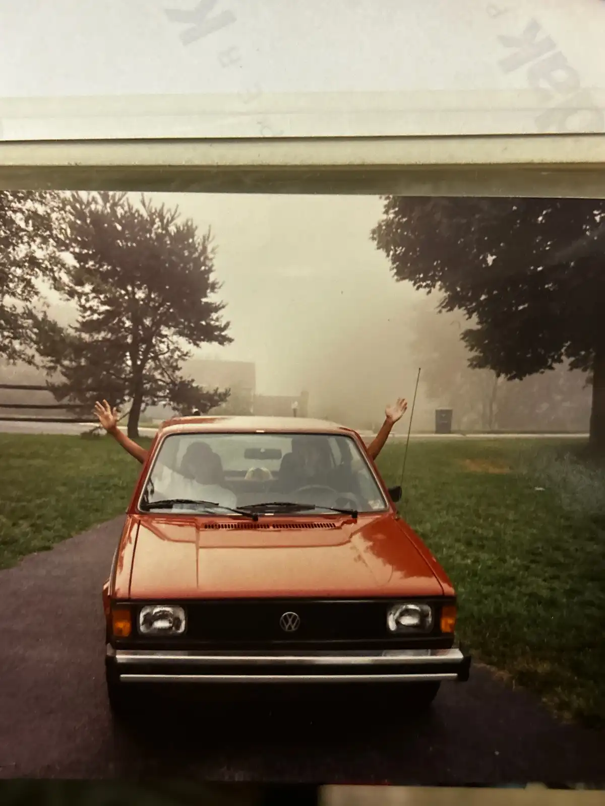 A vintage orange Volkswagen car is parked on a grassy area with trees in the background. Two people are visible inside the…