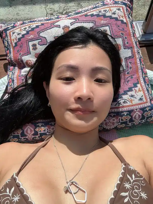 A person with dark hair lies on a patterned pillow outdoors, wearing a brown bikini top with white embroidery and a silver…