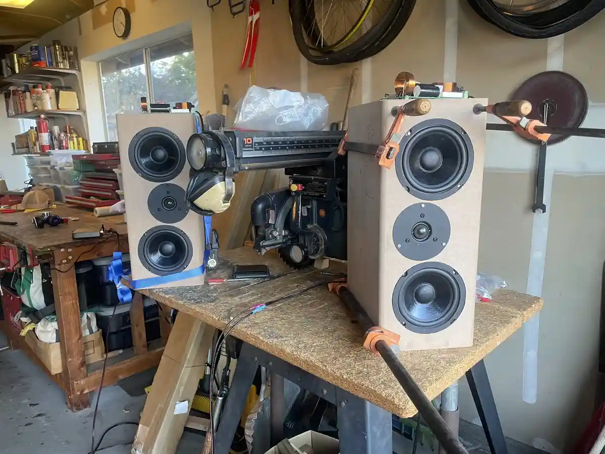 Two homemade speaker cabinets with three drivers each are clamped to a cluttered workbench in a garage, surrounded by tools…