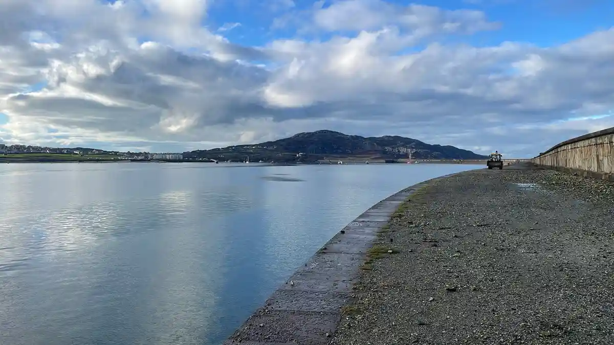 A wide shot of a calm body of water with a gravel path along its edge, leading toward a distant mountain range under a…