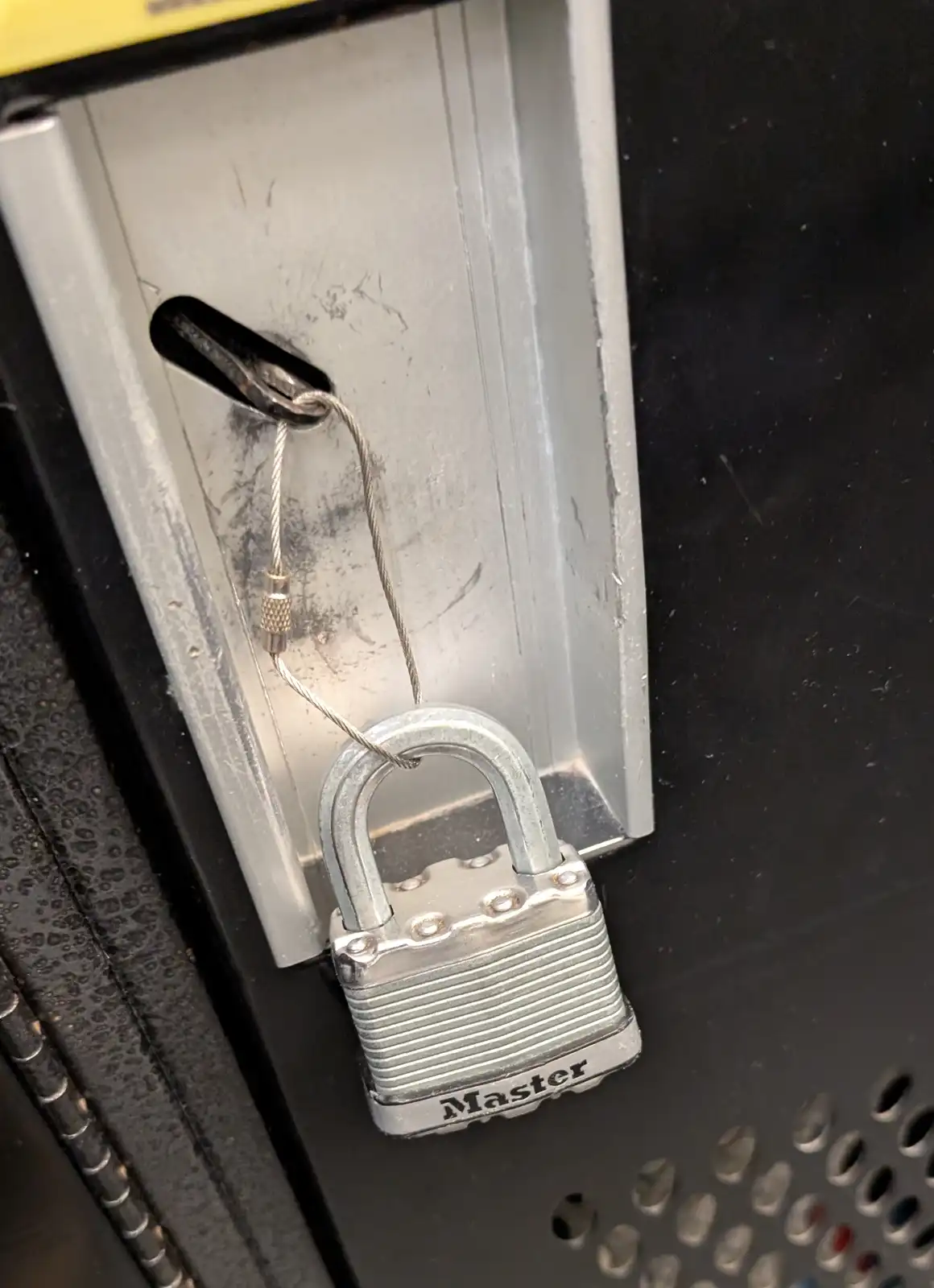 A close-up photo shows a silver Master brand padlock secured with a steel cable through a slot in a metal cabinet door. The…