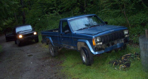 A blue, older model pickup truck is parked on grass next to a dirt road, with a black pickup truck approaching from behind…