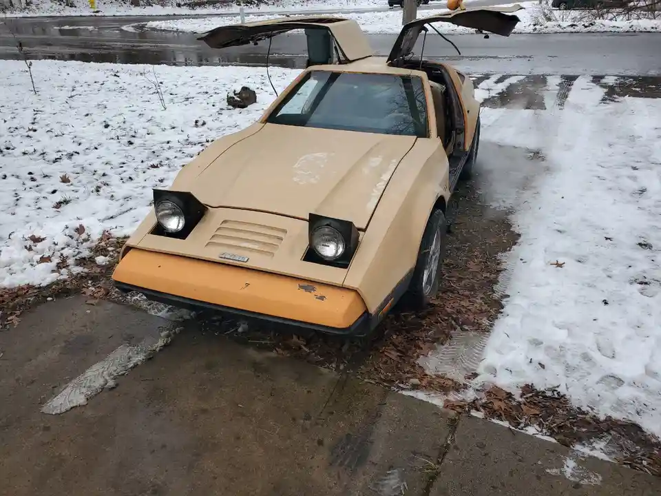 A beige and orange vintage sports car with gull-wing doors open, parked on a snowy driveway with melting snow and leaves…