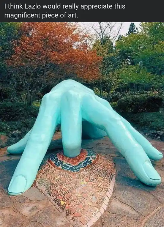 A large, turquoise-colored sculpture of a hand is shown emerging from a mosaic-tiled heart shape on a stone path, surrounded…