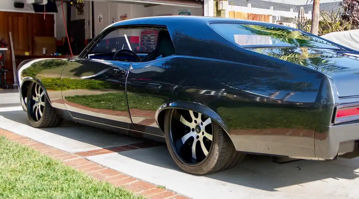 A glossy black classic American muscle car, likely a Chevrolet Chevelle, is parked on a driveway with a brick border and…
