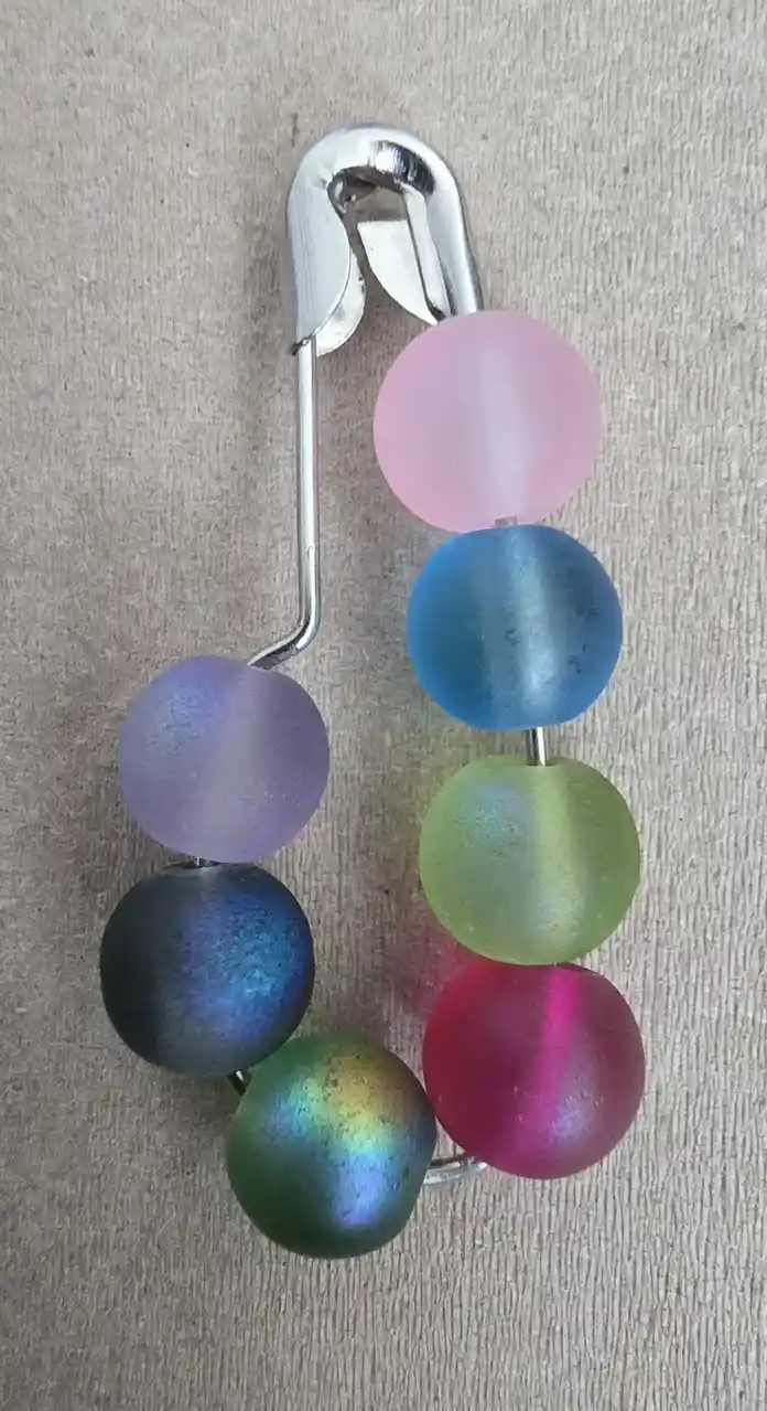 A silver safety pin has several colorful, translucent, spherical beads threaded onto its arms, arranged in a rainbow-like…