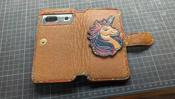 A handmade brown leather phone case with a colorful embroidered unicorn head design sits open on a gray grid cutting mat…