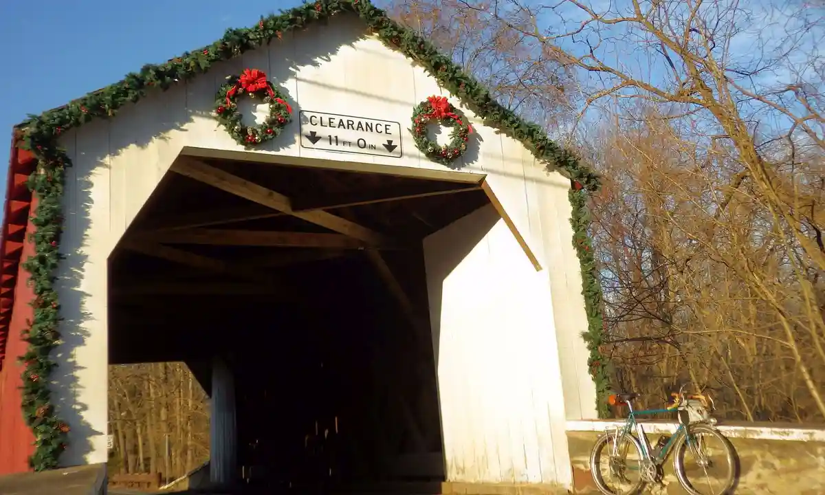 A white wooden covered bridge decorated with green garlands and red bows for the holidays has a sign indicating 11 feet 0…