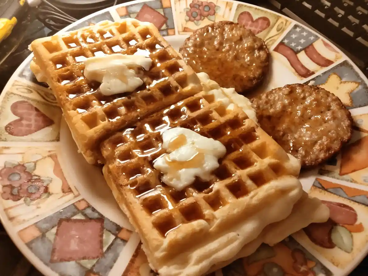 A close-up photo shows a plate of breakfast food including two golden-brown waffles topped with butter and syrup…