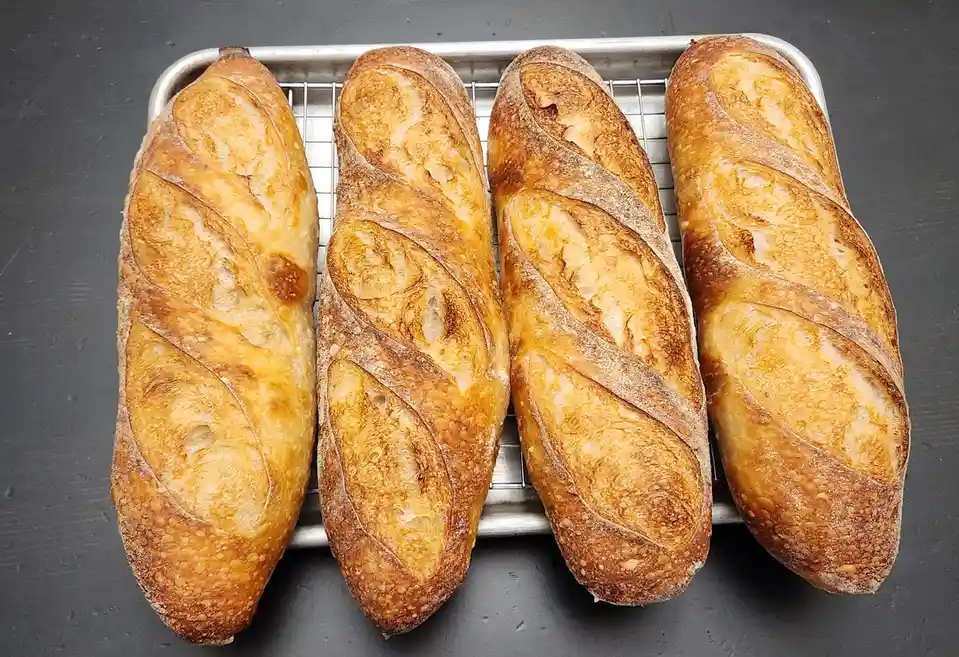 Four golden-brown, crusty baguettes sit side-by-side on a metal cooling rack, resting on a dark gray surface. The bread has…
