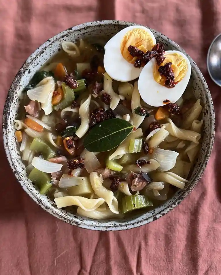 A top-down photo of a rustic ceramic bowl filled with pasta, vegetables, and broth, topped with a halved hard-boiled egg and…
