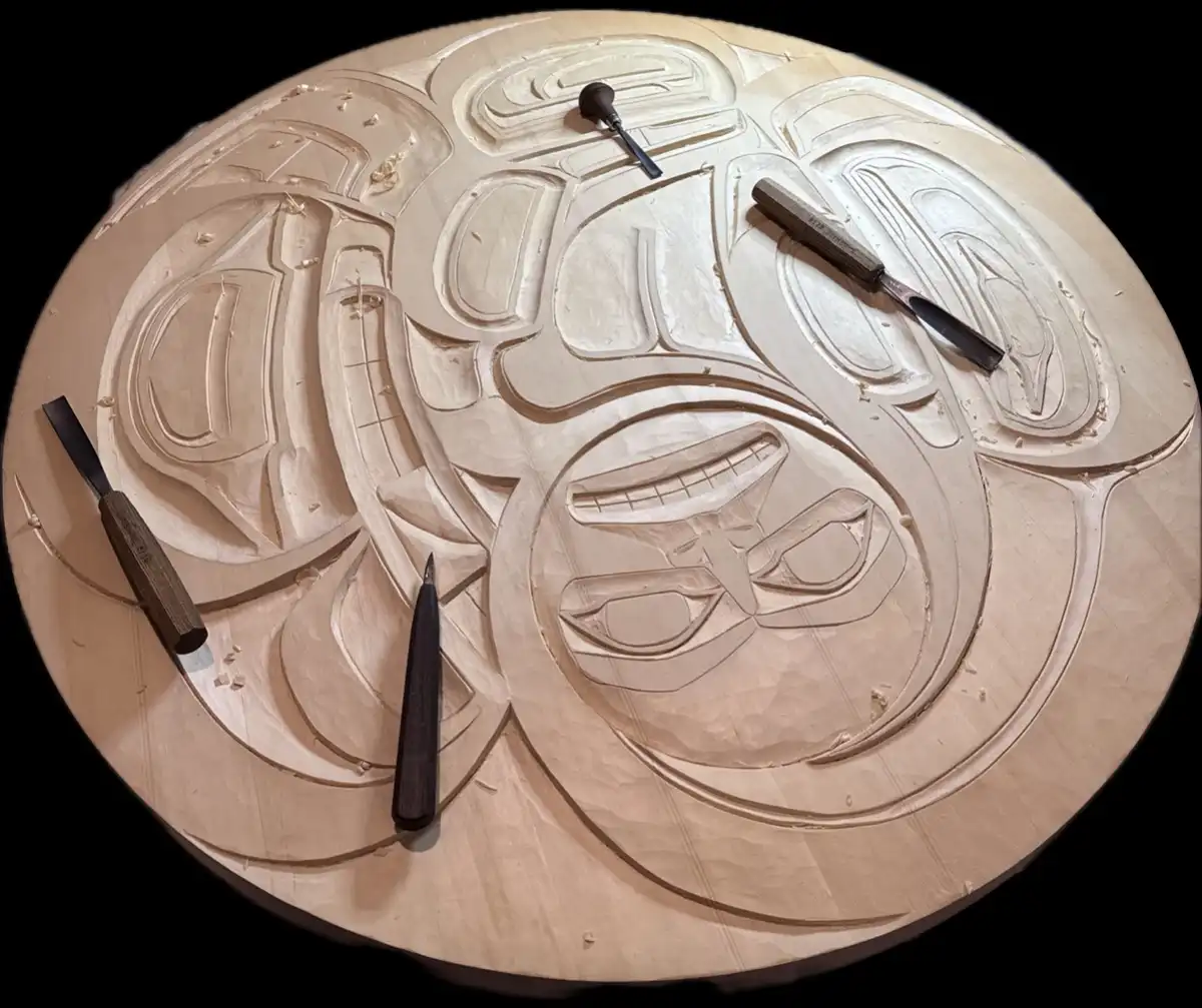 A top-down view of a circular wooden carving in progress, featuring stylized Indigenous Northwest Coast art motifs including…