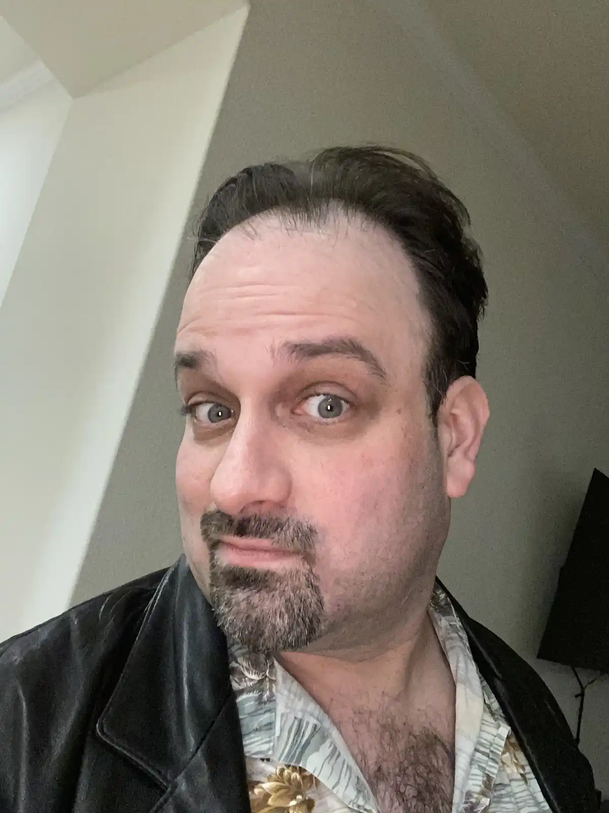 A man with a receding hairline, goatee, and wide-eyed, startled expression stares directly at the camera. He wears a black…
