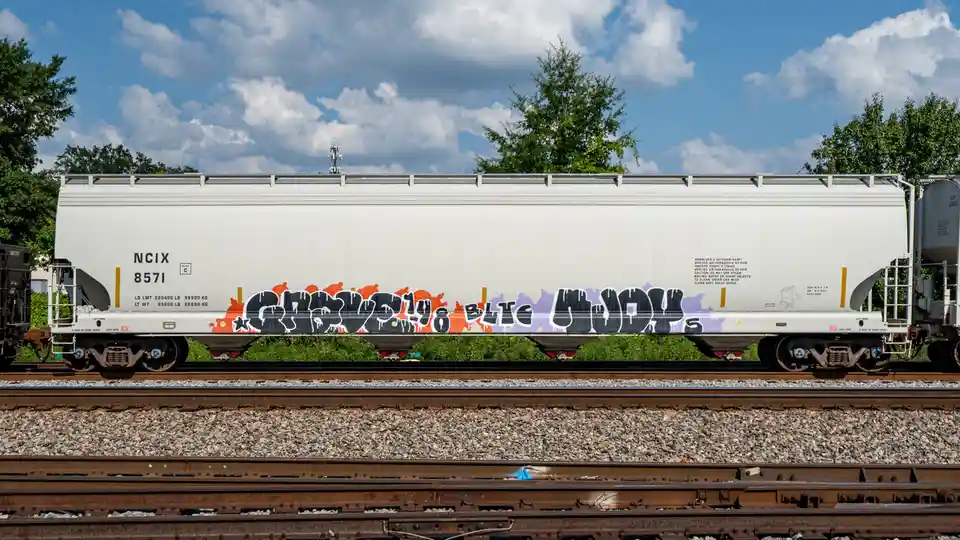 A white freight train car sits on tracks under a partly cloudy sky, featuring large graffiti that reads 'grov3ly8 btc mods'…