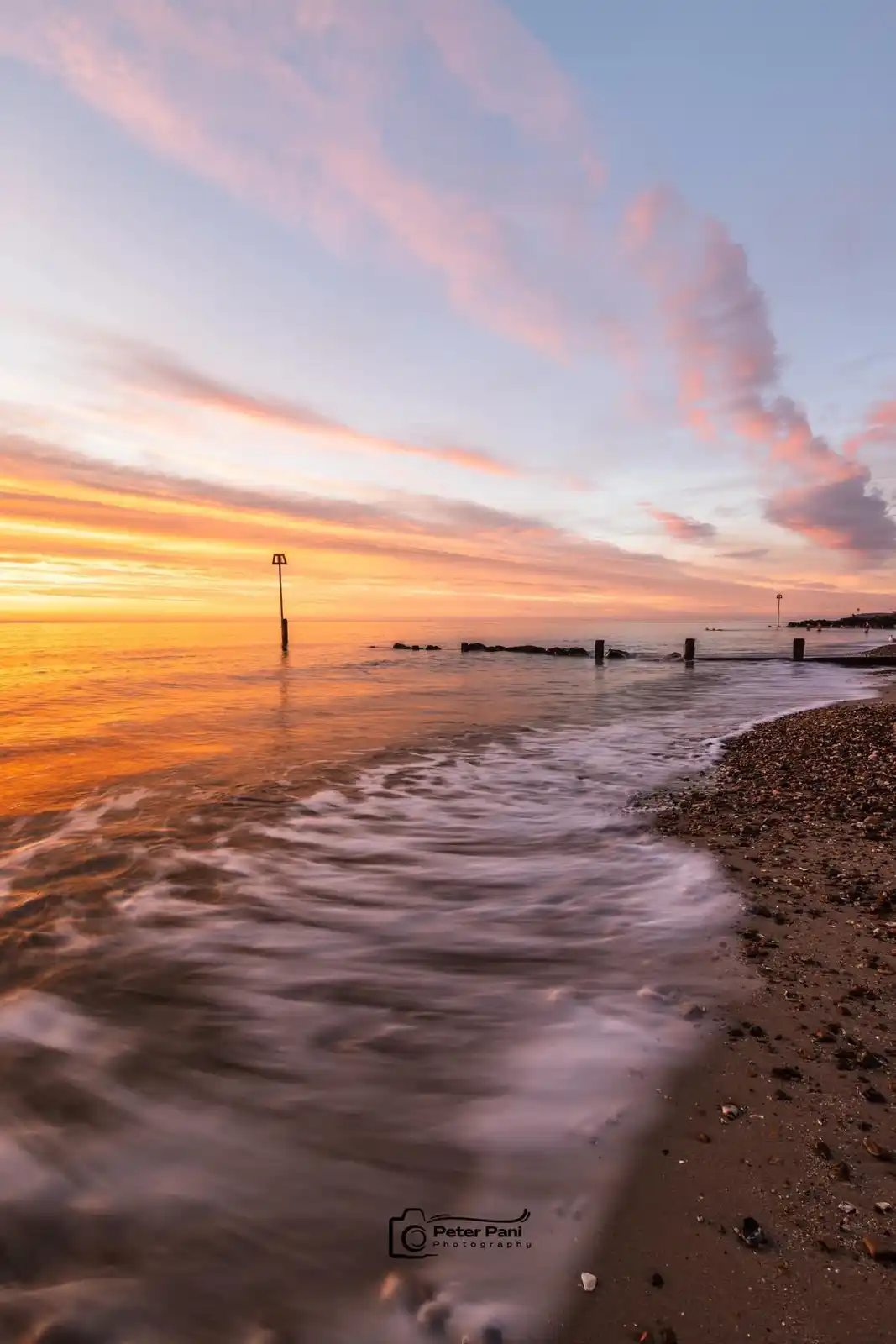 A serene beach scene at sunset with orange and pink clouds reflected in calm water, gentle waves lapping a pebble shore, and…