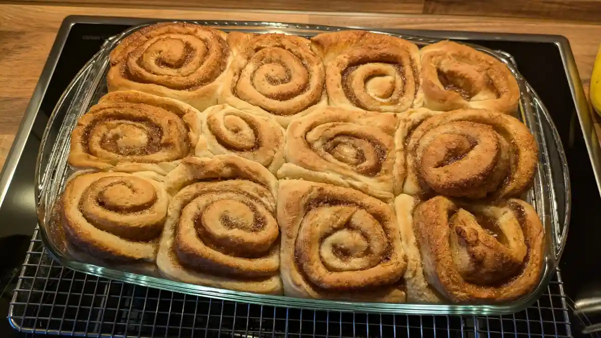 A glass baking dish filled with freshly baked cinnamon rolls sits on a cooling rack atop a black ceramic stovetop. The rolls…