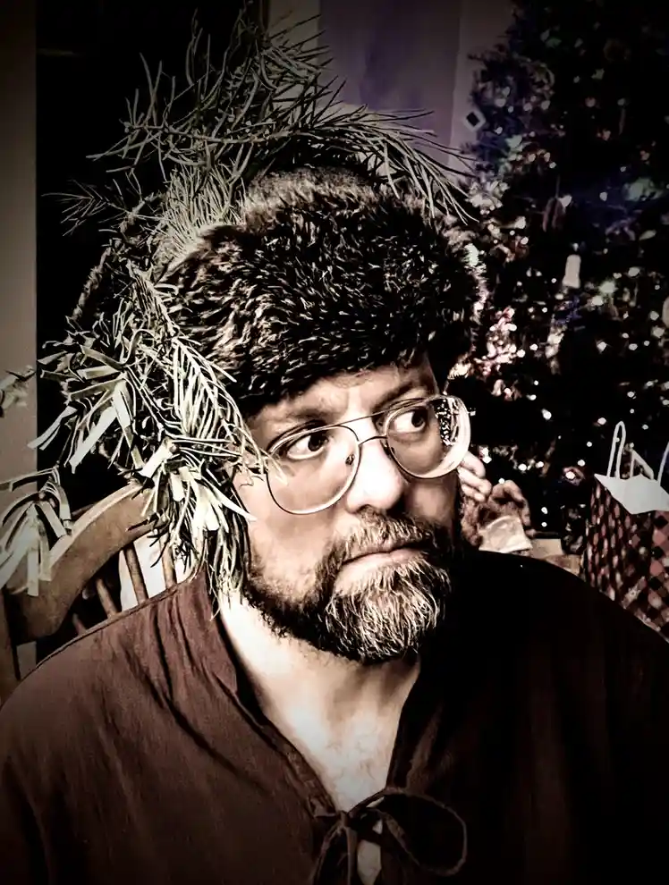 A man with glasses and a beard wears a furry hat with pine branches stuck to it, looking sideways with a weary expression…
