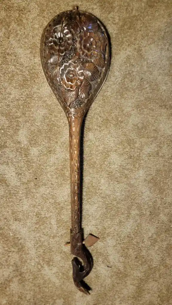 a highly ornate, intricately carved wooden spoon with floral patterns on its bowl and a long handle ending in a hook, lying…