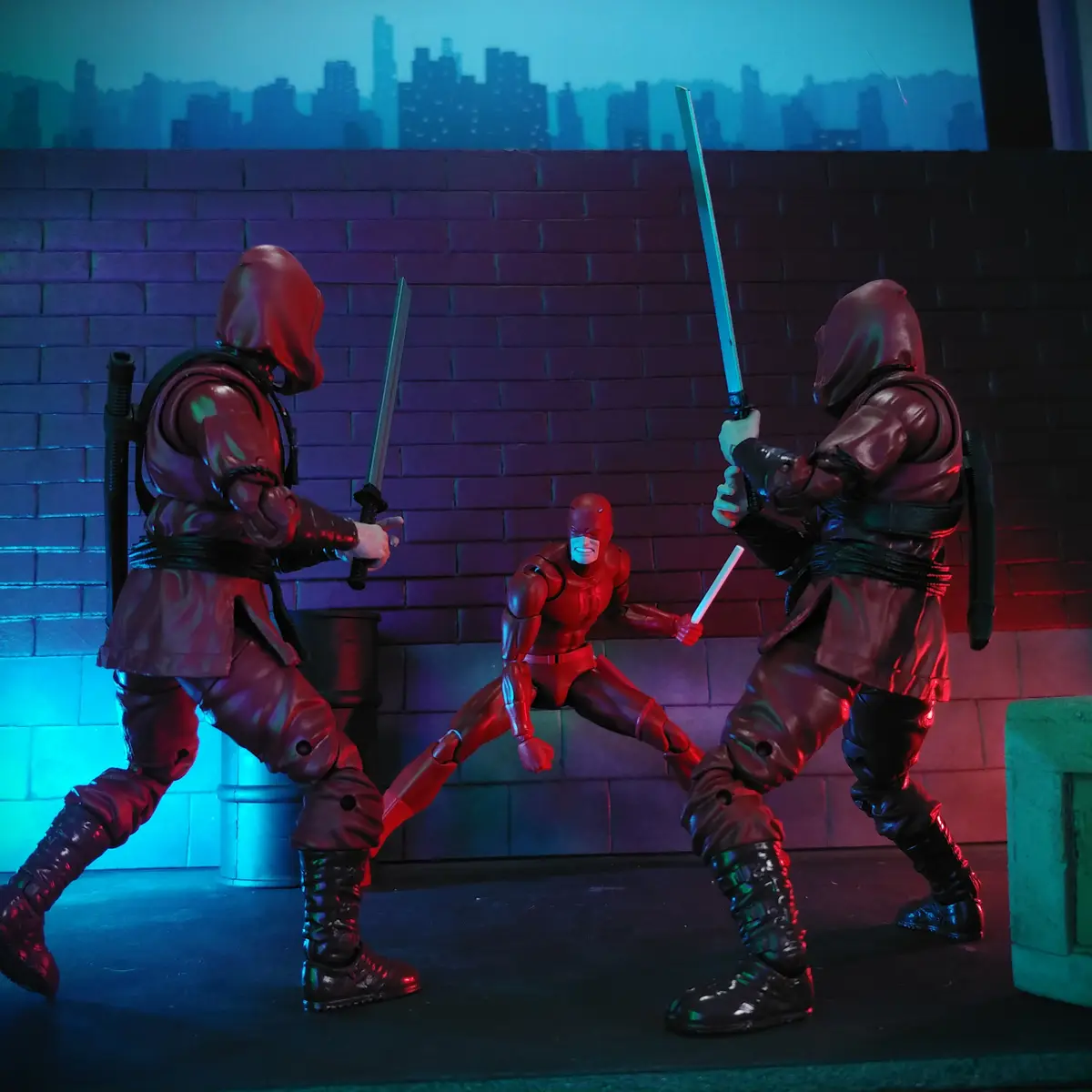 Three action figures are staged in a dramatic pose: Daredevil in red stands between two hooded assassins wielding swords…
