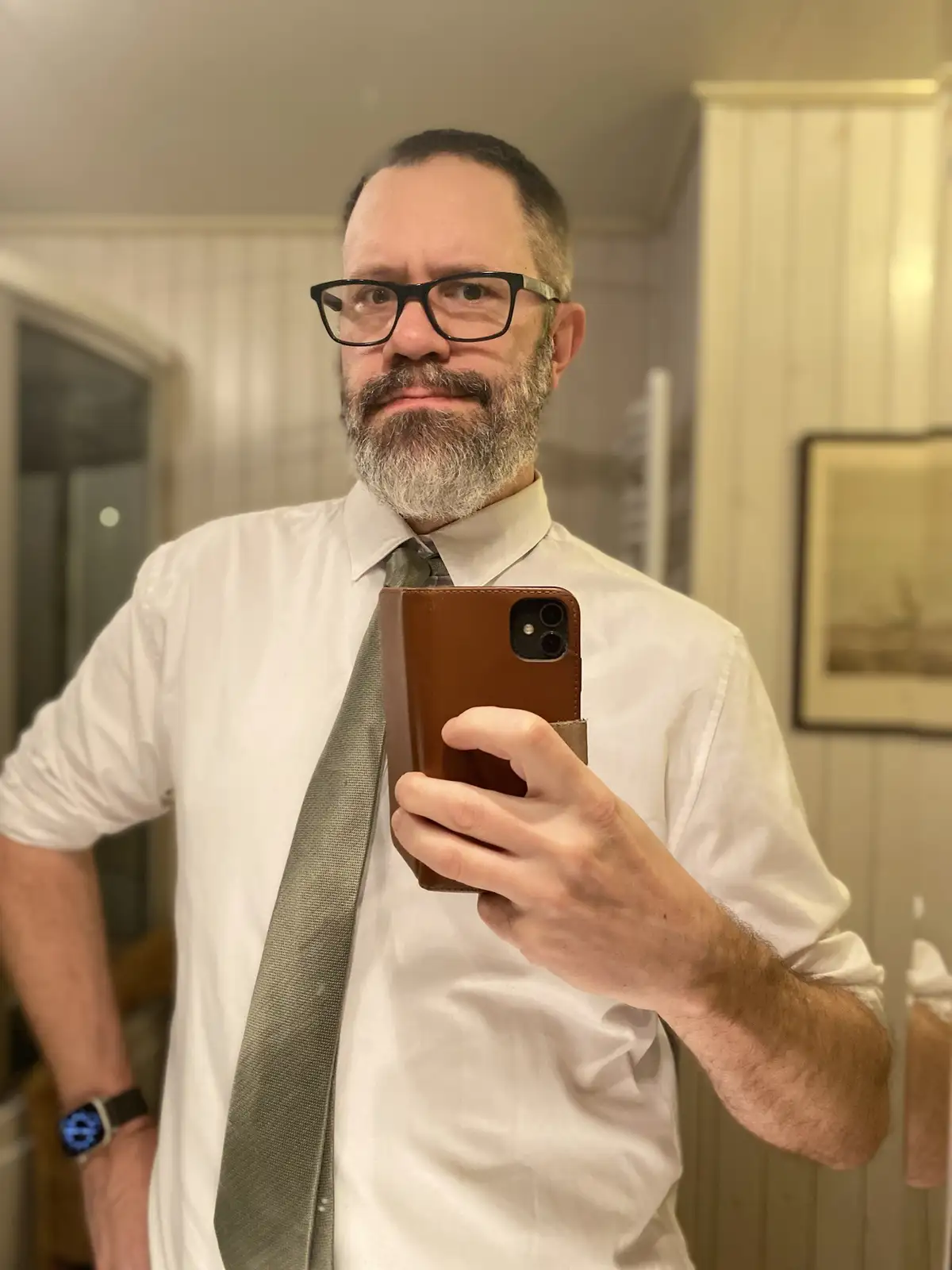 A man with glasses and a gray beard takes a mirror selfie in a white shirt and gray tie, holding a phone in a brown case. He…