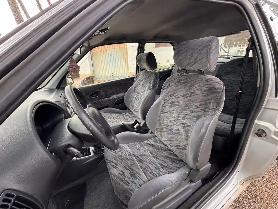 The image shows the interior of a car with worn, patterned gray fabric seats, a black steering wheel, and a visible seatbelt…