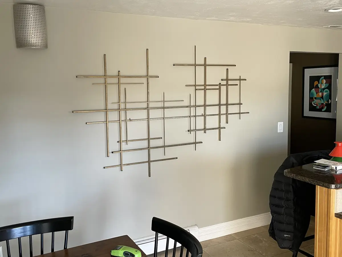 A photo of a modern dining room wall with abstract metal wall art made of intersecting rods. The art is arranged in two…