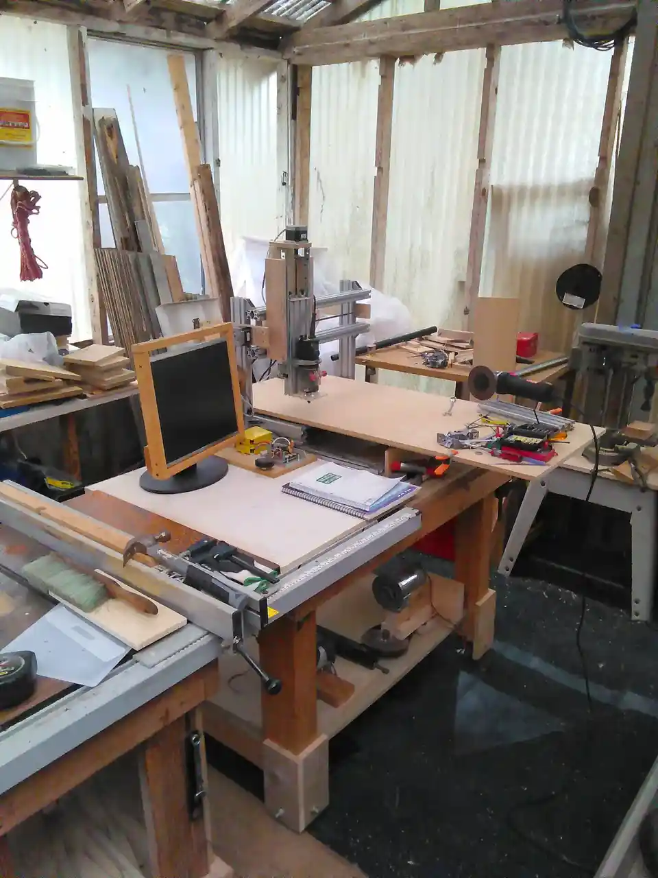 A cluttered DIY workshop features a homemade CNC router mounted on a workbench, with a small monitor nearby. The text…