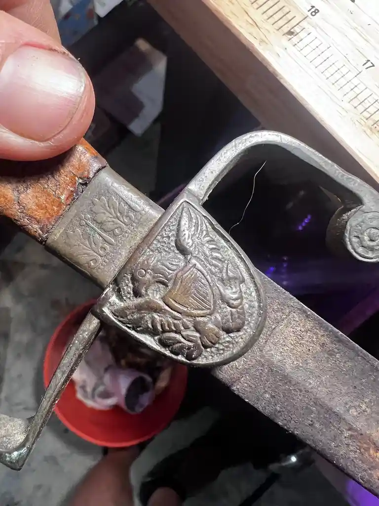 A close-up photo shows a hand holding an old, ornate sword with a wooden grip and metal hilt. The pommel features a detailed…