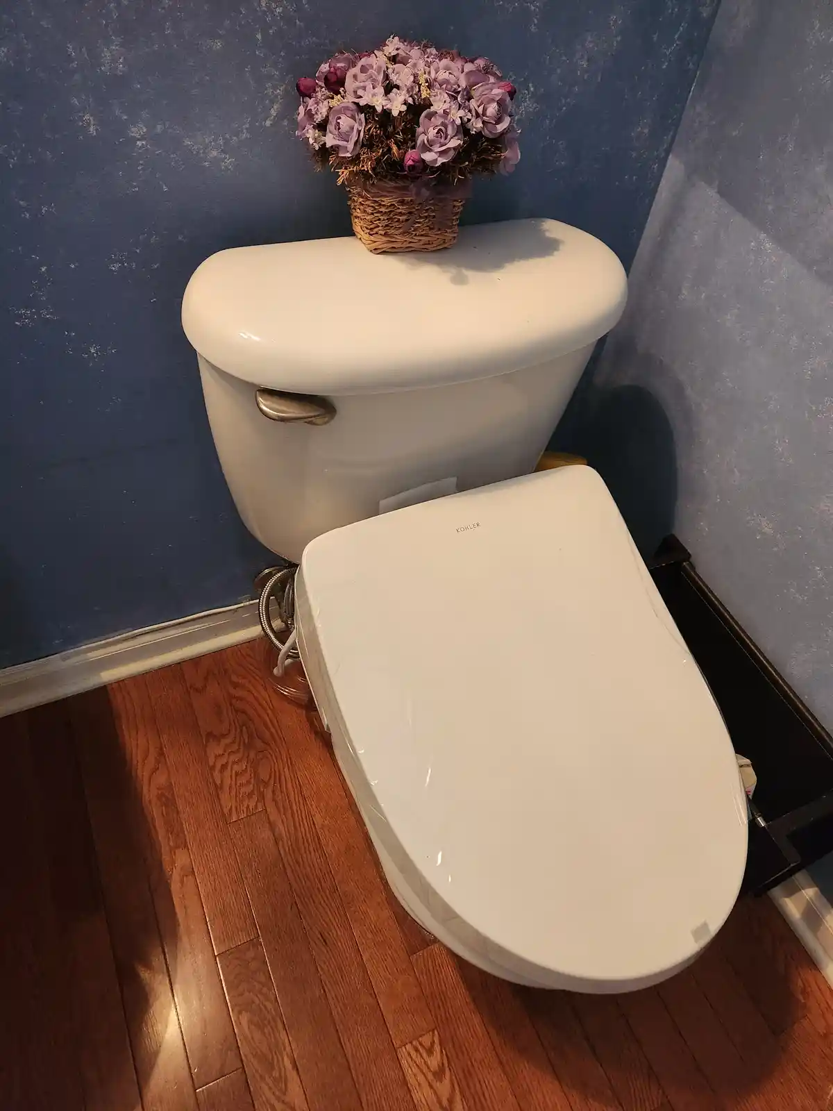 A white Kohler toilet sits in a bathroom with blue textured walls and wooden flooring. A woven basket filled with purple and…