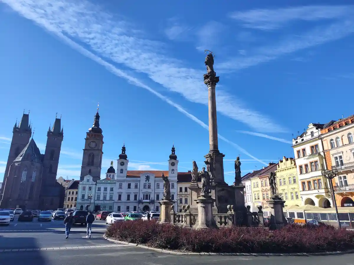 A bright, sunny photo of a European city square featuring a tall stone column monument with statues, surrounded by colorful…