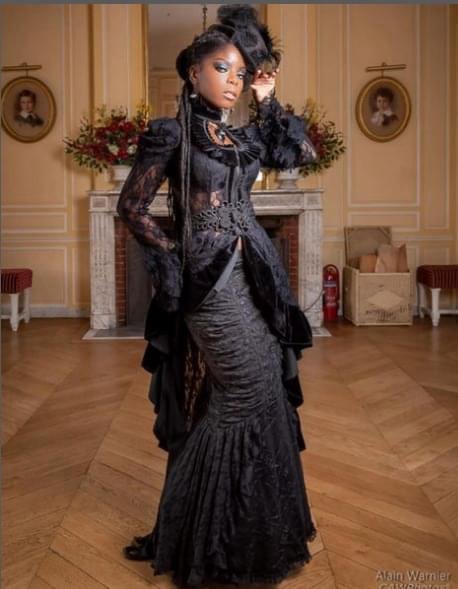 A Black woman in an elaborate, ornate black Victorian-style gown with lace, ruffles, and a feathered hat poses confidently…