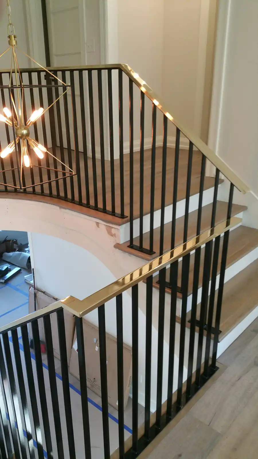 A modern staircase with black metal balusters and gold-colored handrails, viewed from above. A contemporary gold chandelier…
