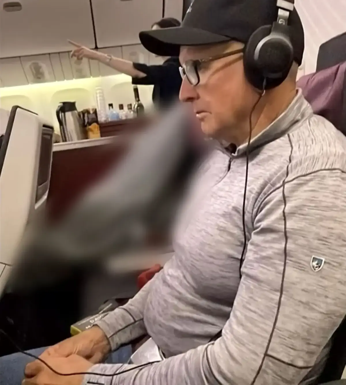 A man wearing glasses, a black cap, and over-ear headphones sits in an airplane seat, looking away with a neutral…