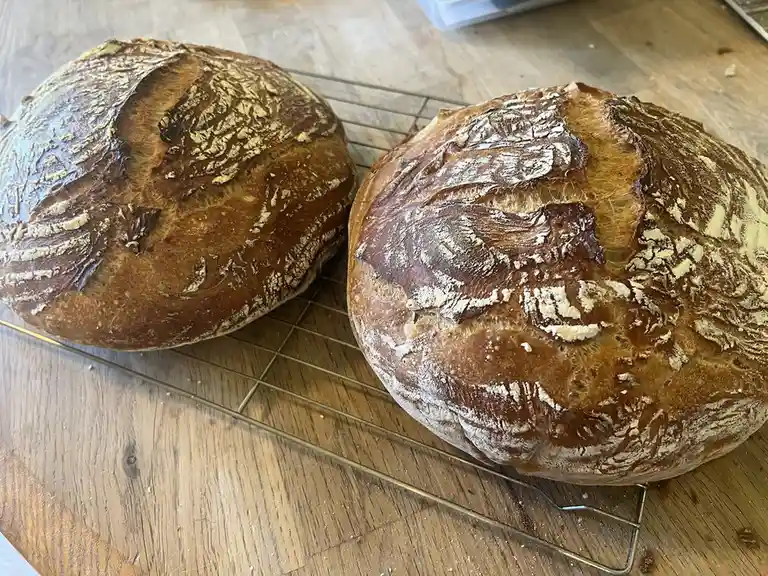 Two round, rustic loaves of bread with golden-brown crusts and flour dusting sit on a wire cooling rack atop a wooden…