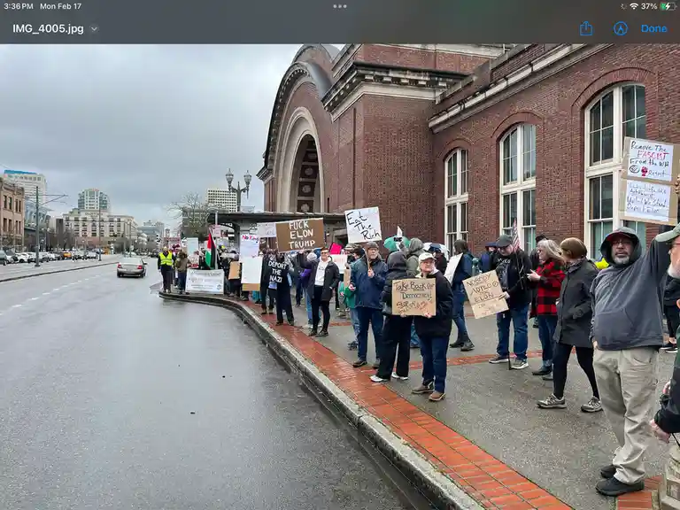 A group of protesters stands on a wet sidewalk in front of a large brick building, holding handmade signs with messages like…