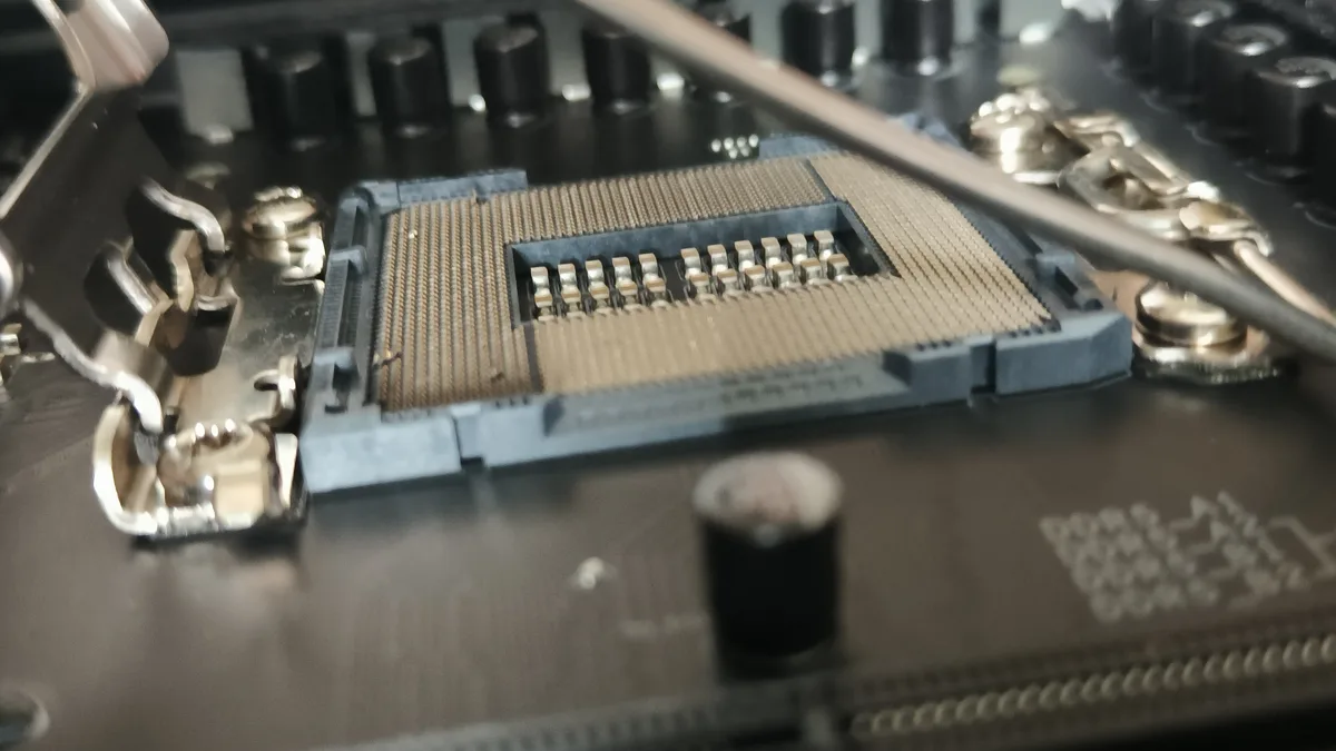 A close-up photo of an empty CPU socket on a motherboard, showing the gold pins and surrounding components. The overlaid…