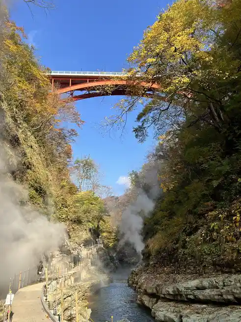 a scenic view of a canyon with a red arched bridge spanning above, surrounded by autumn foliage in yellow and green, with…