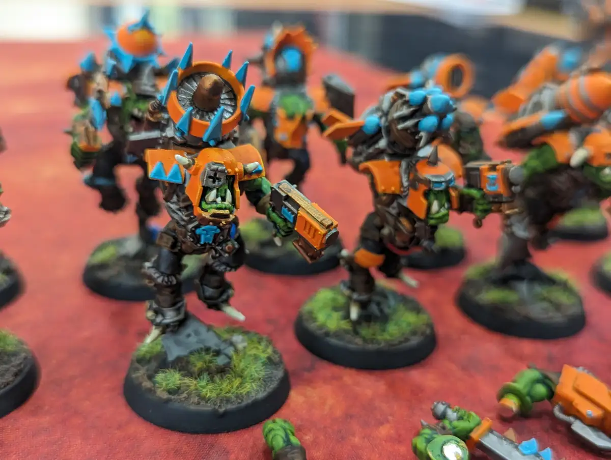A close-up photo shows a group of painted miniature orc warriors in orange and blue armor, standing on circular bases with…