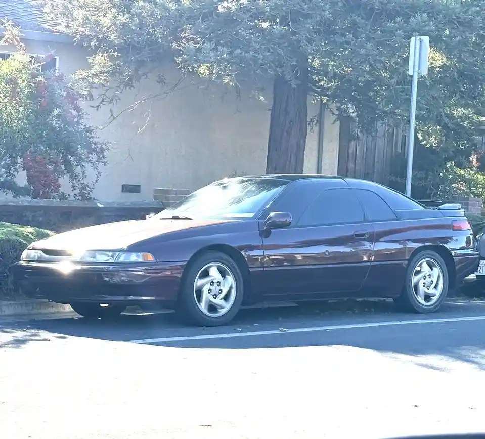 A maroon 1990s-era coupe, likely a Toyota Celica or similar, is parked on a residential street with trees and a house in the…