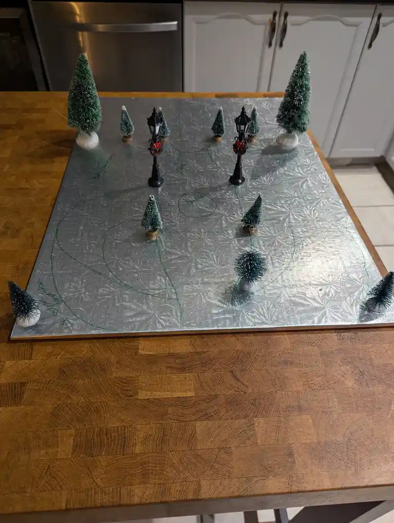 A homemade tabletop hockey rink made from a silver foil-covered board on a wooden table, featuring miniature Christmas trees…