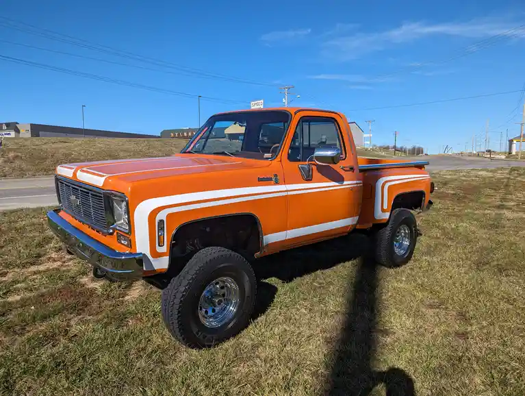 A bright orange classic Chevrolet pickup truck with white racing stripes is parked on grass beside a road under a clear blue…