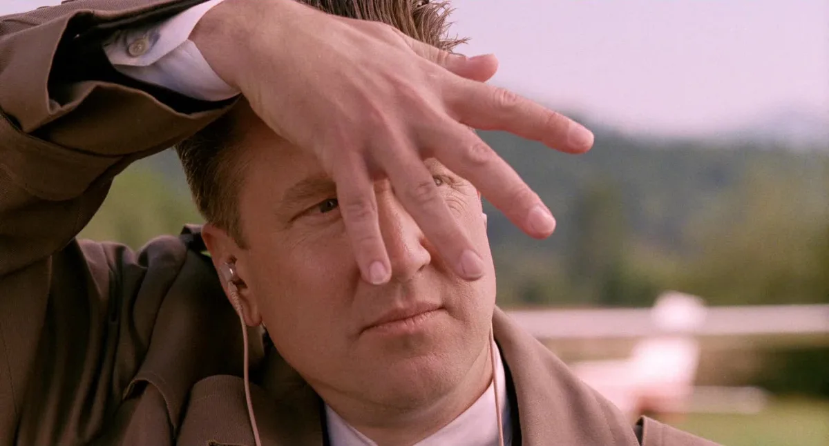 a man in a brown jacket and white shirt has his hand over his face, partially obscuring his eyes, while wearing earphones…