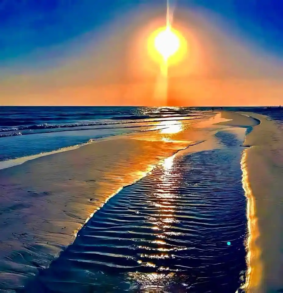 A vibrant beach sunset with the sun casting a bright, vertical beam of light down to the water's edge. The sand is wet and…