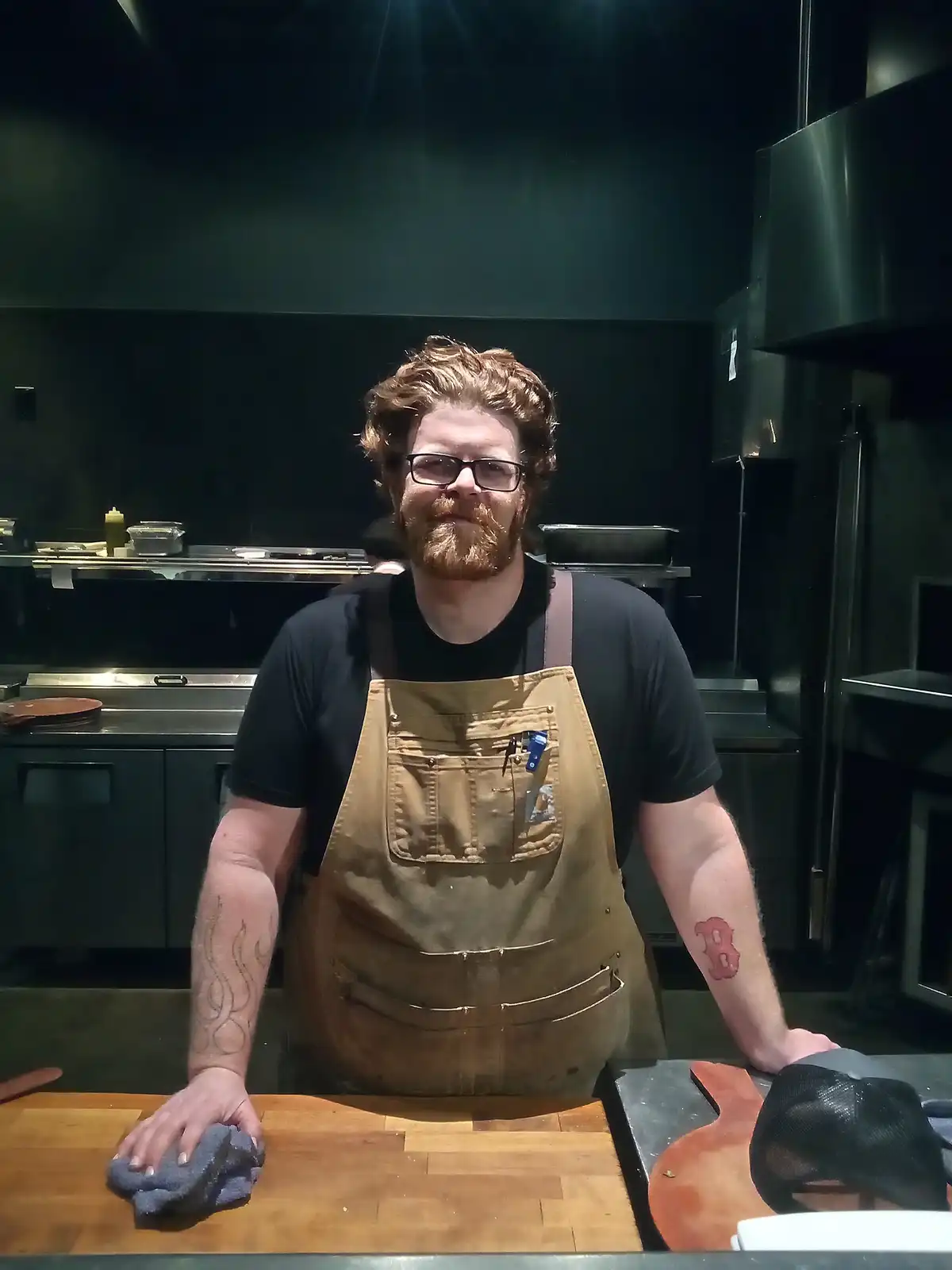 A man with a reddish beard and glasses, wearing a black t-shirt and a brown work apron, stands in a commercial kitchen. He…