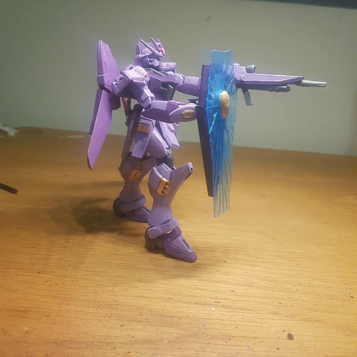 A purple Gundam-style robot model stands on a wooden table, holding a blue translucent shield and a rifle. The robot has…