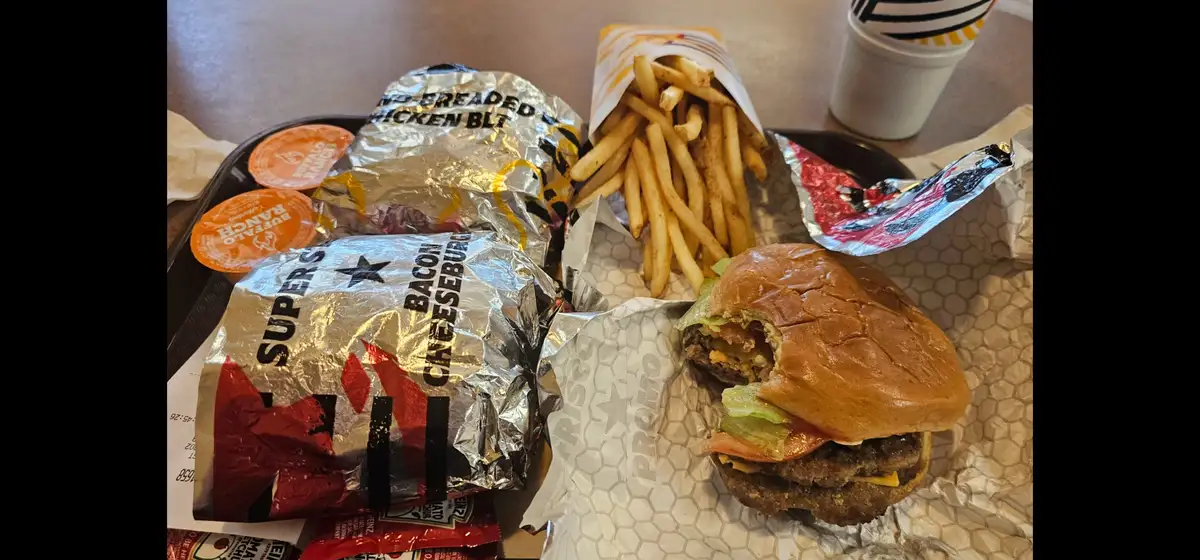 A close-up photo of a fast-food meal on a tray, featuring a partially eaten bacon cheeseburger, a bag of fries, and two…